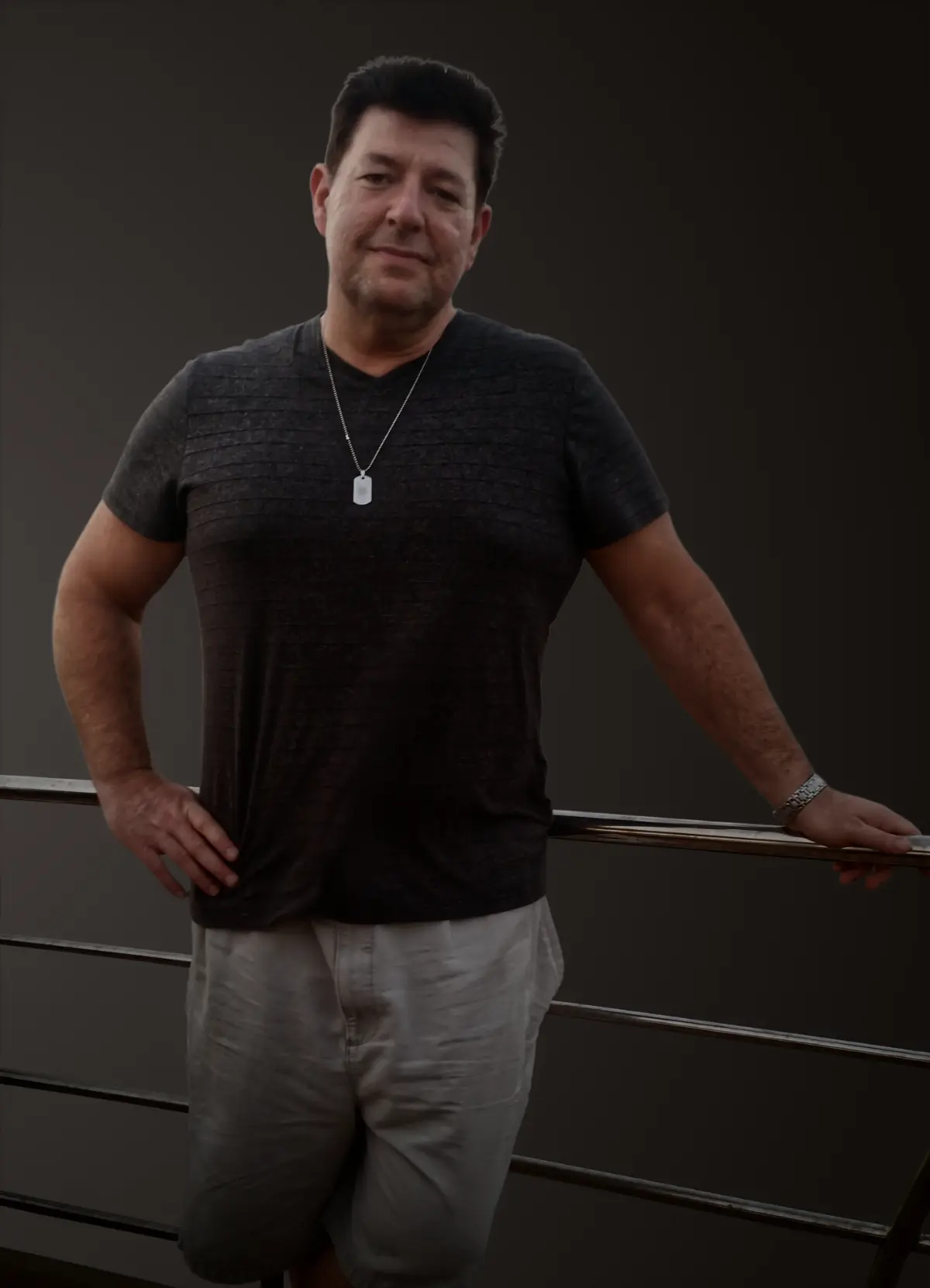 A man wearing a dark t-shirt, light shorts, and a necklace stands by a metal railing against a plain dark background.